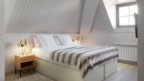 Une chambre avec un plafond en pente, un lit recouvert d’une couverture à rayures, deux lampes de chevet, une table de chevet et une fenêtre avec des stores.