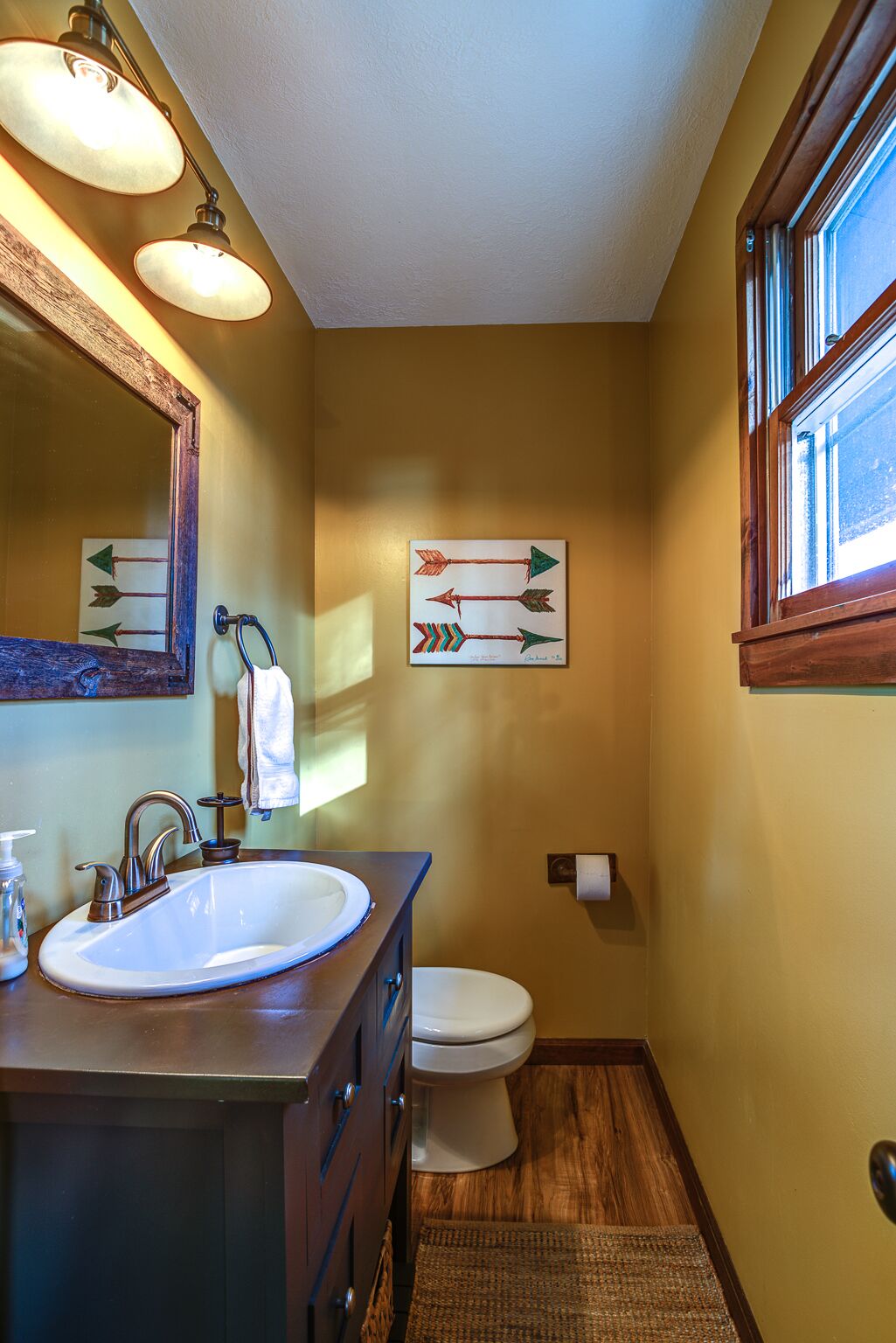 Une salle de bain avec un lavabo, un miroir et des toilettes. Des flèches sont peintes sur le mur et il y a une fenêtre avec des stores.