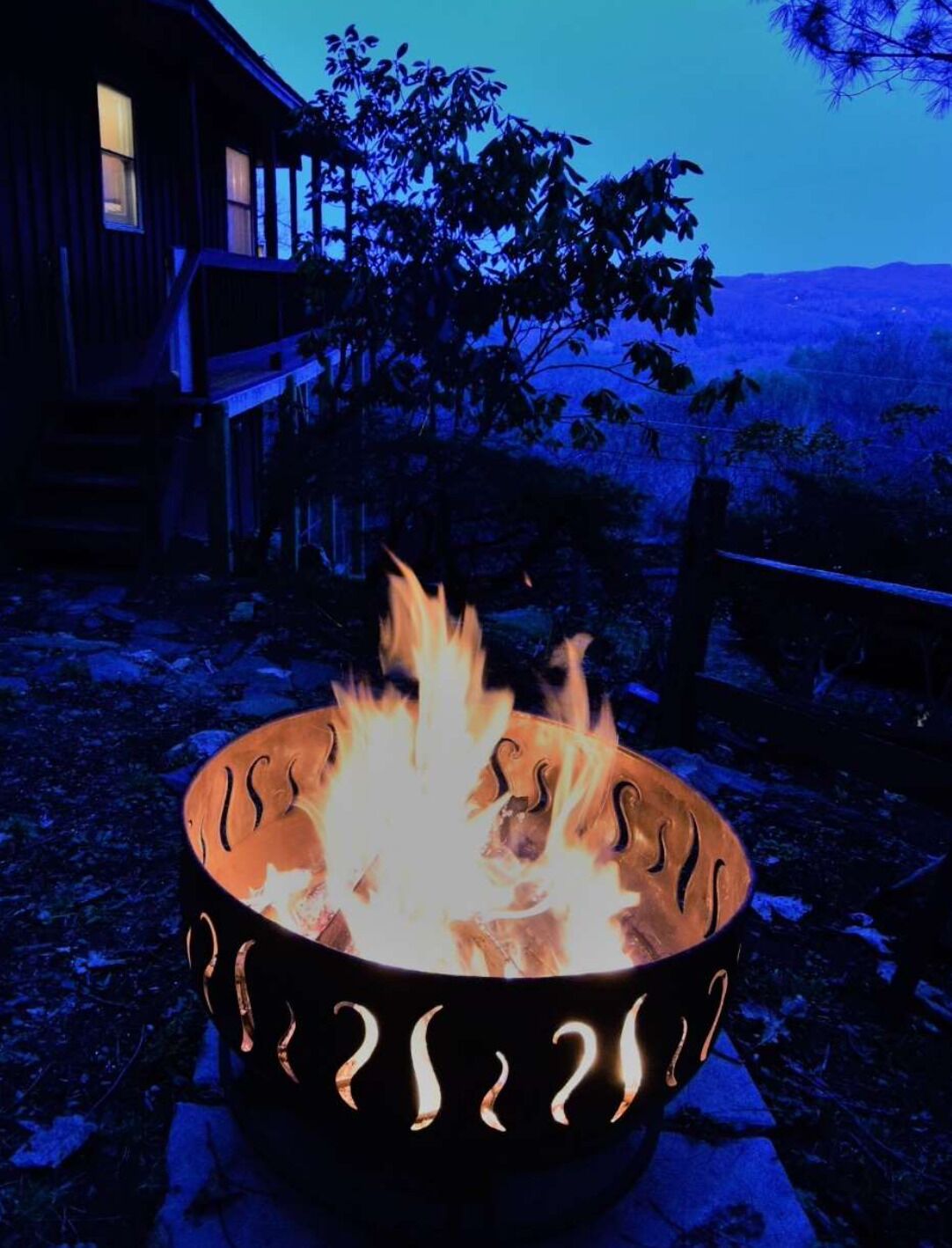 Un foyer avec des flammes, entouré d’une terrasse en bois et d’une maison en arrière-plan.