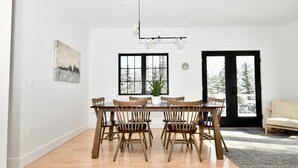 A dining area with a wooden table and chairs, a chandelier, a window with a view of trees, and a door leading outside.