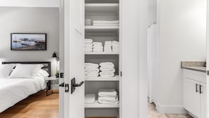 A hotel room with a neatly organized closet containing folded towels, a bed with white bedding, a bedside table with a lamp, and a framed picture on the wall.
