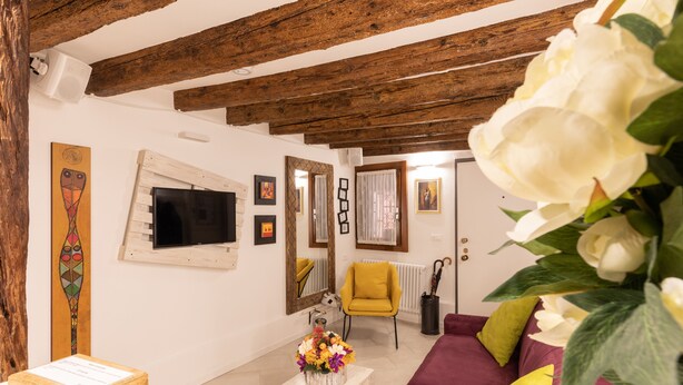 A living room with exposed wooden beams, a television, a yellow armchair, and a sofa with colorful cushions. - “Ea casa de mì - The Magic of Experiencing Venice” (Venezia)