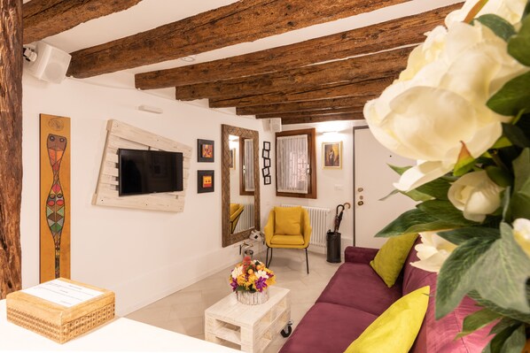 Una sala de estar con vigas de madera a la vista, un televisor, un sillón amarillo y un sofá con cojines de colores.