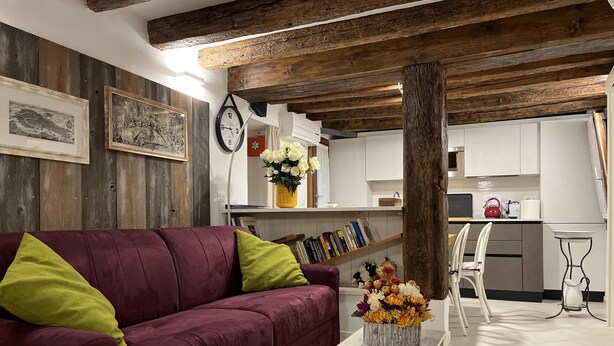 A living room with a purple sofa, a wooden coffee table with a floral arrangement, a clock, and a rustic ceiling. - “Ea casa de mì - The Magic of Experiencing Venice” (Venezia)
