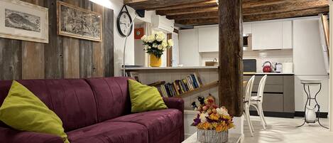 Un salon avec un canapé violet, une table basse en bois agrémentée d’un arrangement floral, une horloge et un plafond rustique.