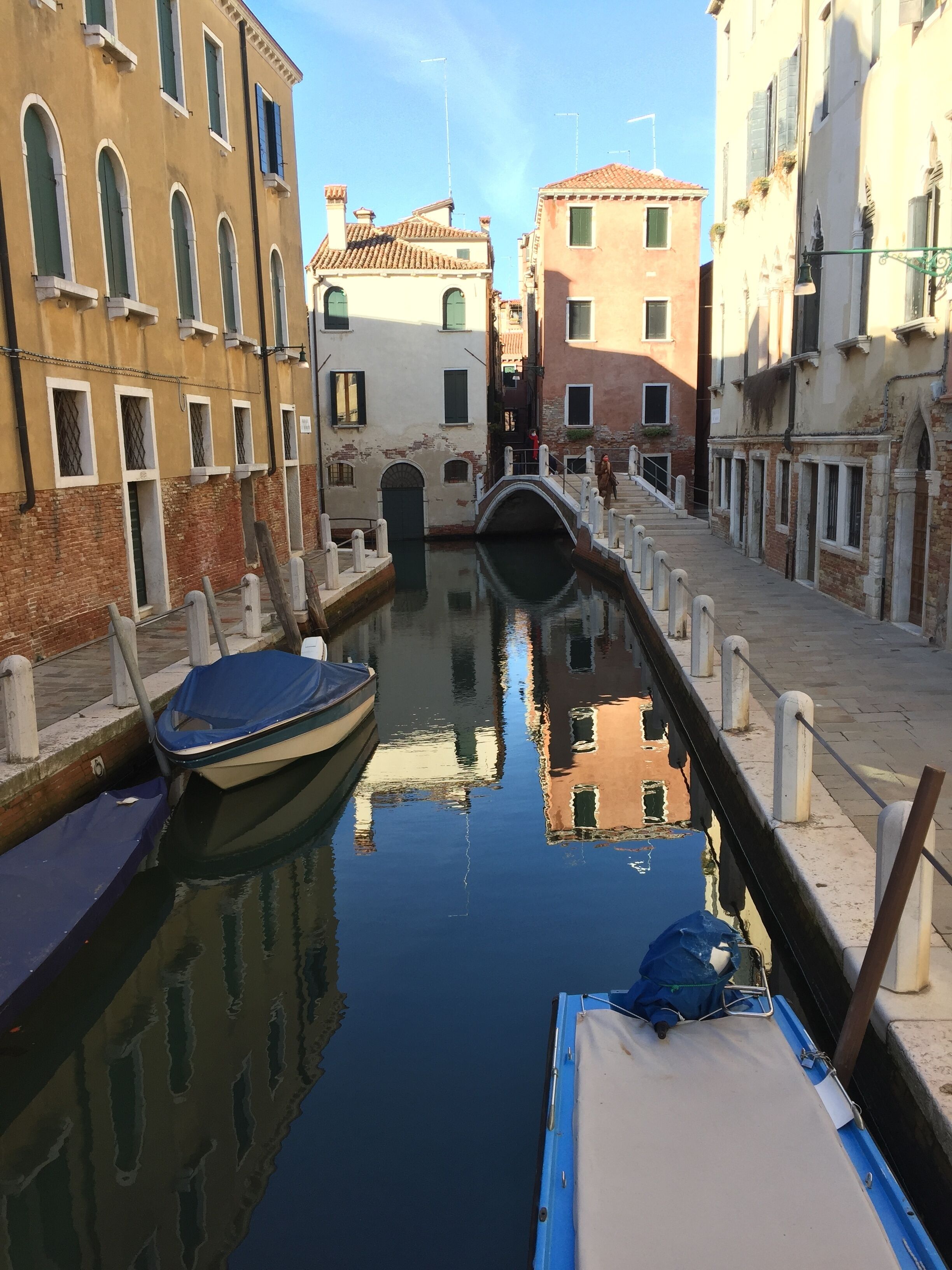 A narrow canal with boats docked on either side, flanked by historic buildings with arched windows and a bridge in the distance.
