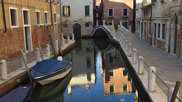 A narrow canal with boats docked on either side, flanked by historic buildings with arched windows and a bridge in the distance. - “Ea casa de mì - The Magic of Experiencing Venice” (Venezia)