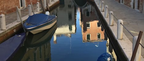 A narrow canal with boats docked on either side, flanked by historic buildings with arched windows and a bridge in the distance.