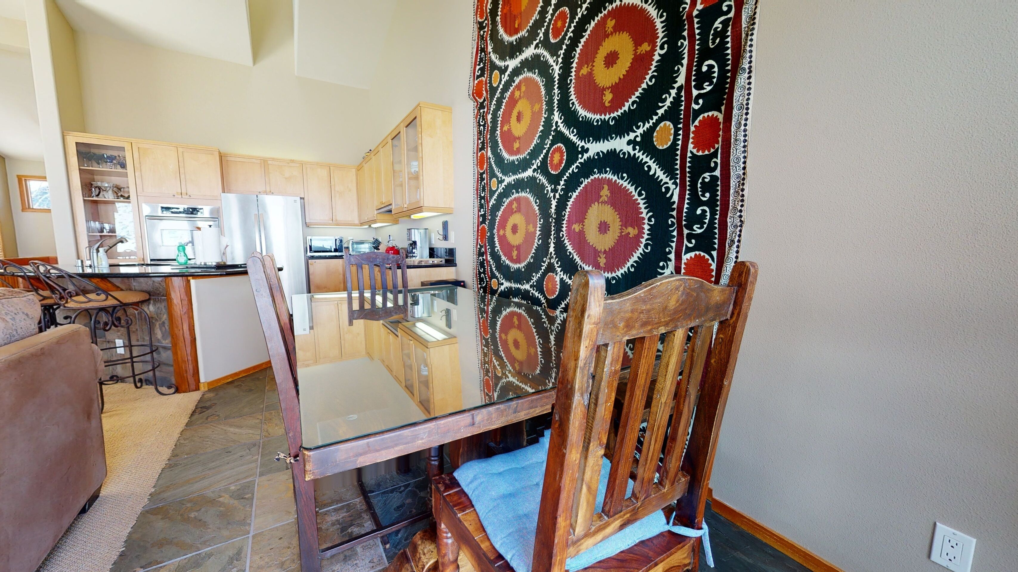 A dining area with a glass table and wooden chairs, a kitchen with wooden cabinets, and a patterned curtain.