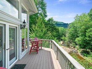 A wooden deck with a table and chairs, overlooking a river and forested hills. - Riverfront Sanctuary | Your Mountain Getaway (Burnsville)