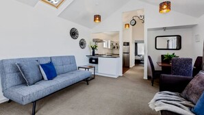 Un salon moderne avec un canapé gris, un coin repas avec une table et des chaises, une cuisine en arrière-plan et des œuvres d’art décoratives au mur.