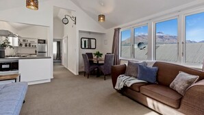 Un salon moderne avec un canapé marron, un coin repas avec des chaises, une cuisine équipée d’un réfrigérateur et de grandes fenêtres offrant une vue sur les montagnes.