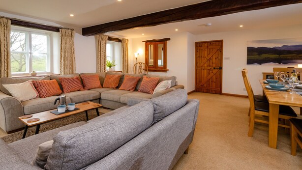 A cozy living room with a large sectional sofa, a wooden dining table with chairs, and a view of the countryside through the windows. - Barn End - Three Bedroom House, Sleeps 6 (Skelwith)