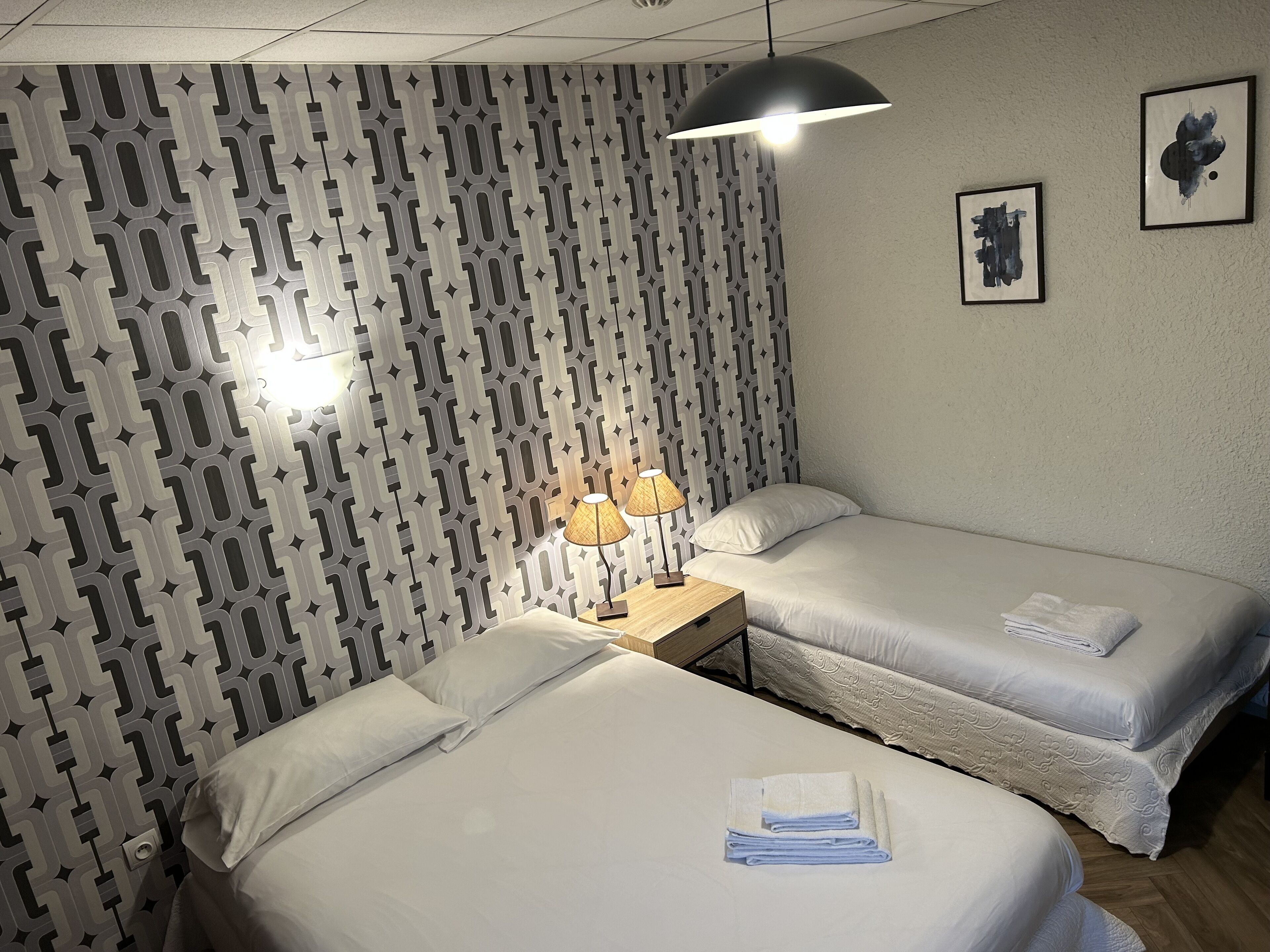 A hotel room with two single beds, a patterned wall, a hanging lamp, and two framed pictures on the wall.