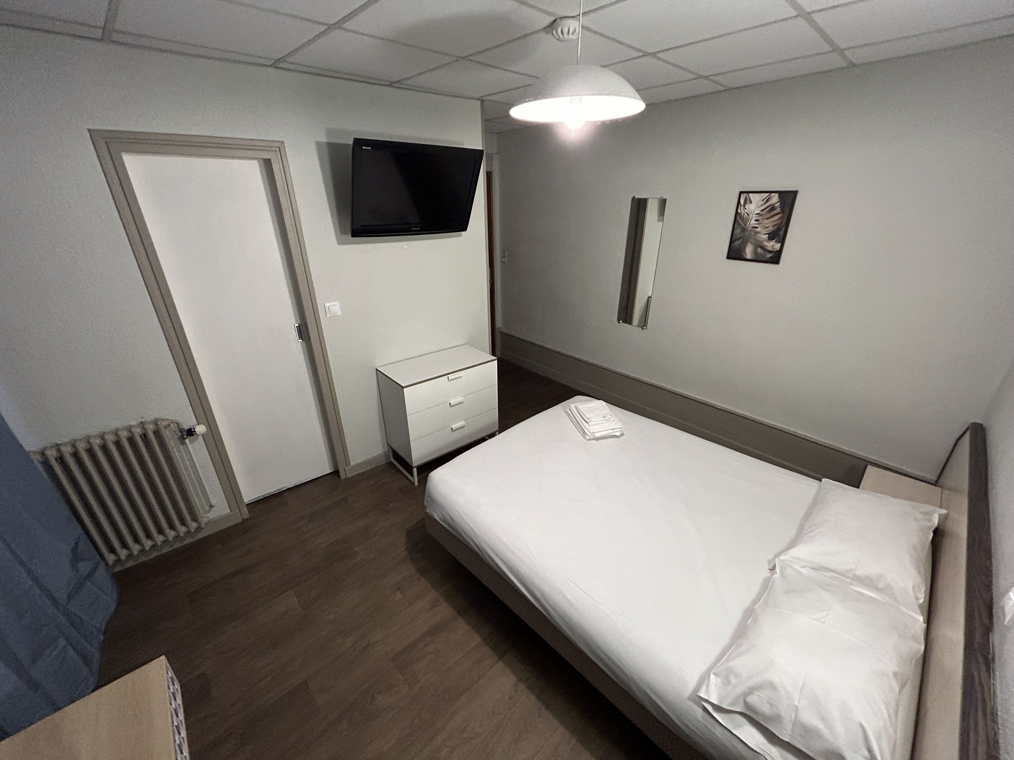 Una habitación de hotel con cama, televisor montado en la pared, mesita de noche y radiador.