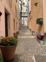 Uma rua de paralelepípedos estreita, ladeada por edifícios com varandas, plantas em vasos e uma caixa de correio verde.
