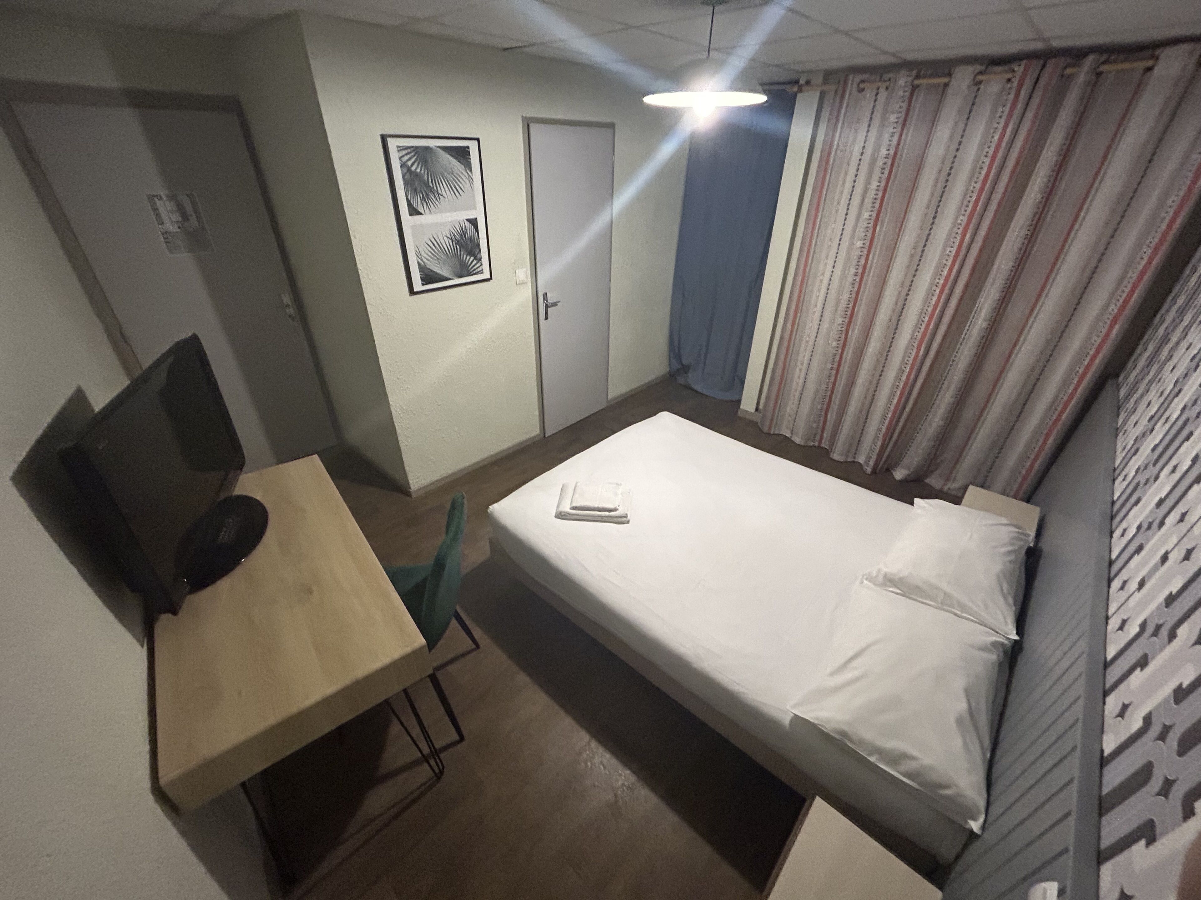 A small, single-room hotel room with a bed, a desk with a computer, a chair, a door, and a window with curtains.