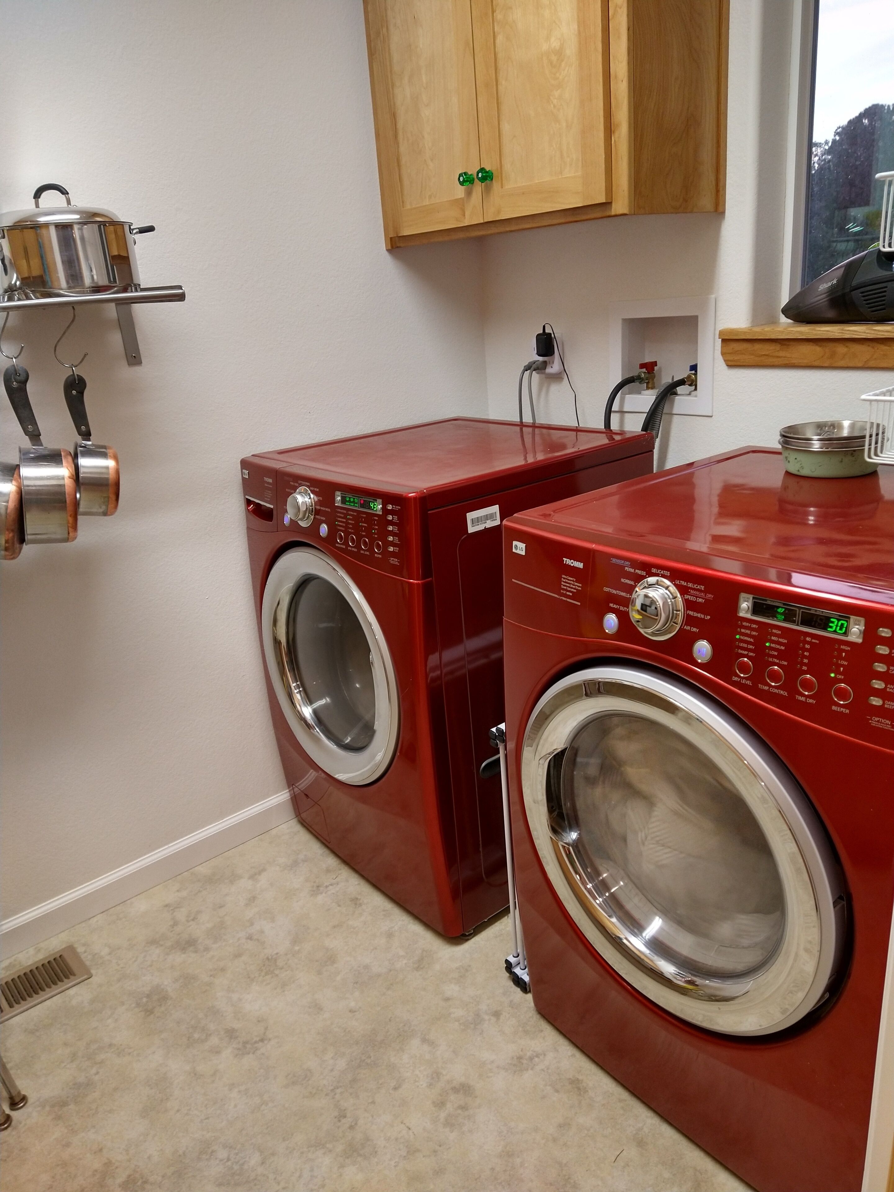 A laundry room with two red front-loading washing machines, a wooden cabinet above, and a metal shelf with pots.