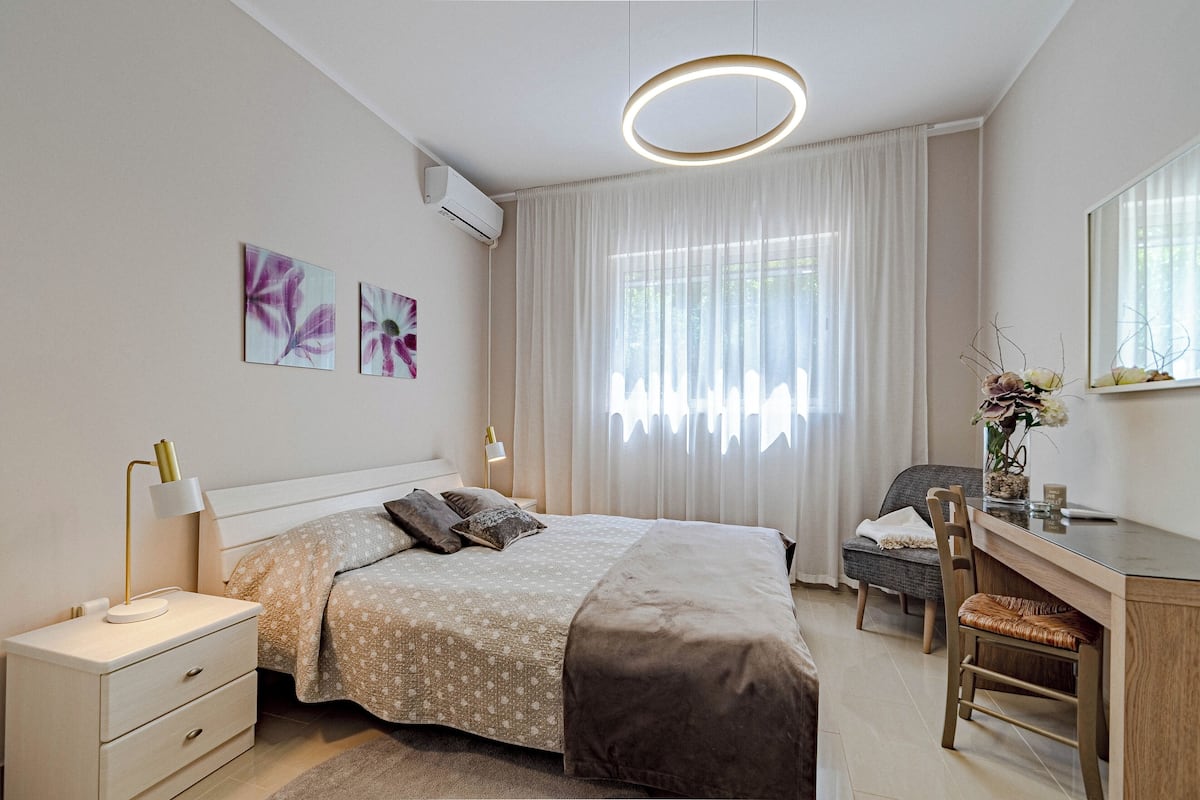Een slaapkamer met een bed, nachtkastje, lamp en een bureau met stoel.