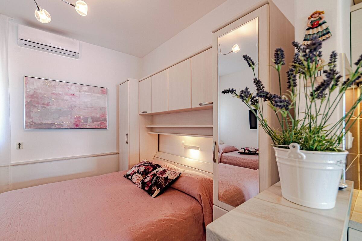Een slaapkamer met een bed, nachtkastje, spiegel en een bloempot.