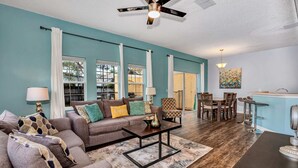 A living room with a sofa, coffee table, and dining area with chairs. There is a ceiling fan, a lamp, and a painting on the wall. - Fresh Decor With Hardwood Floors Huge Private Pool Area CDC Standards 4tv121 (Kissimmee)