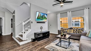 A modern living room with a staircase, a flat-screen TV, a ceiling fan, and a large window with curtains. - Fresh Decor With Hardwood Floors Huge Private Pool Area CDC Standards 4tv121 (Kissimmee)