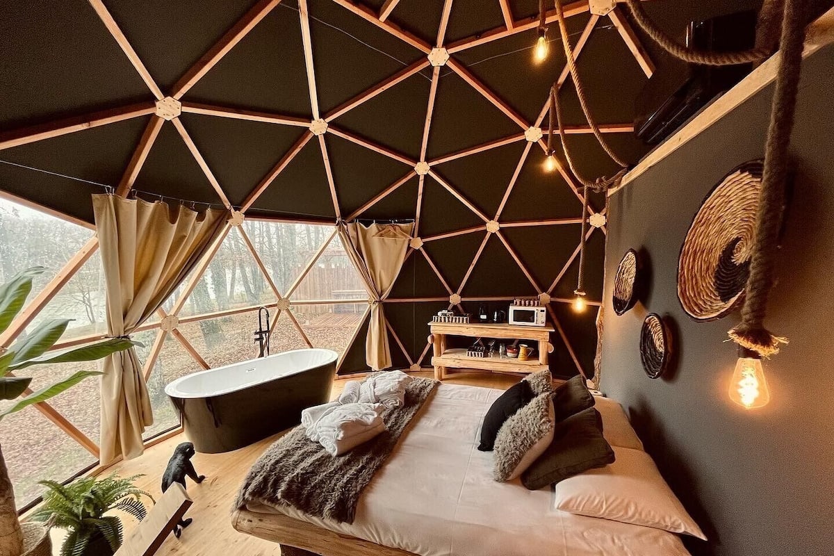 A cozy bedroom with a large bed, a wooden bench, a bathtub, and a small wooden shelf with items.