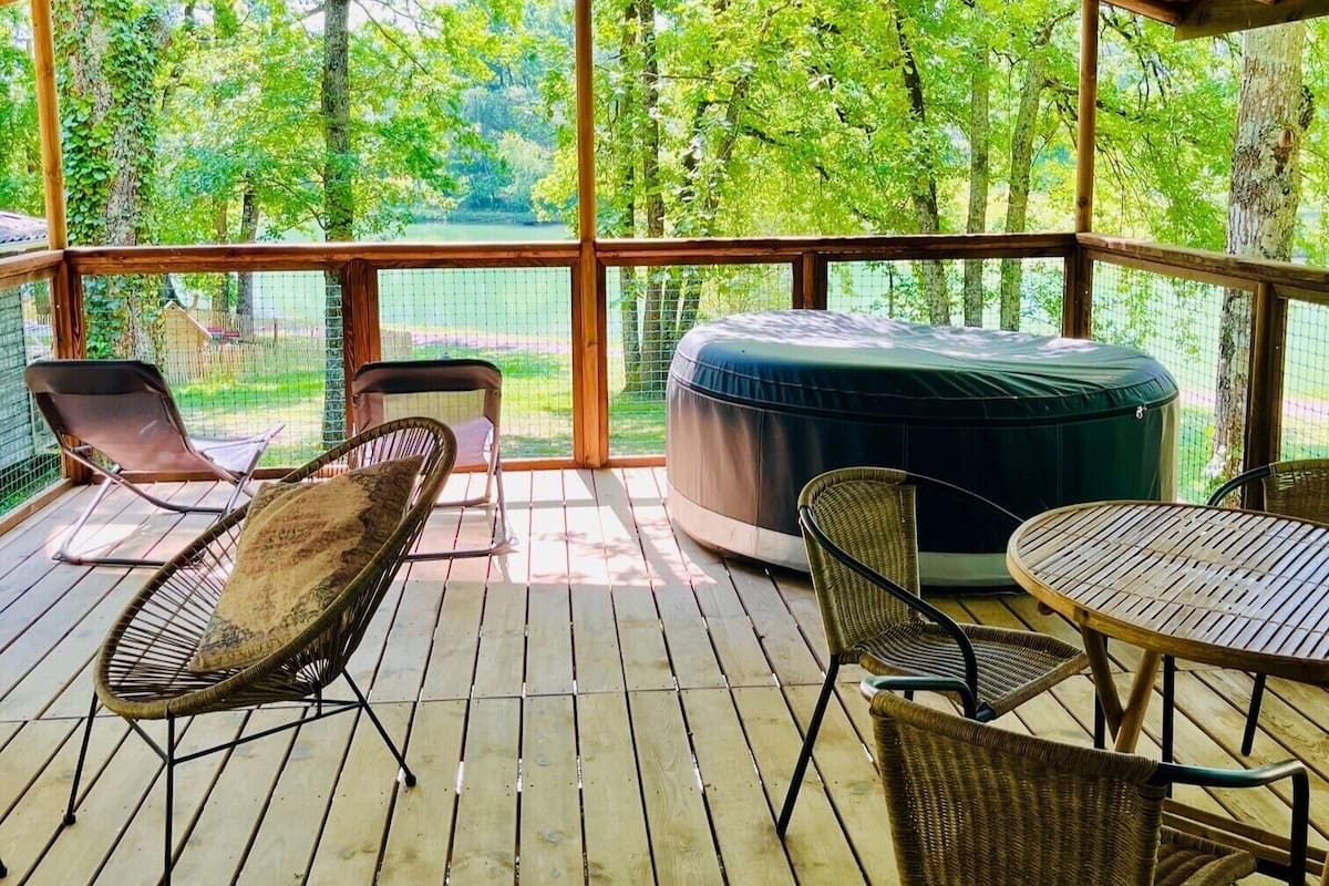 A covered outdoor area with wicker chairs, a round table, and a cushioned seating area, overlooking a natural landscape.