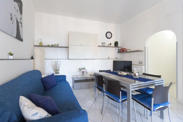 Una sala de estar moderna con un sofá azul, una mesa de comedor con sillas, un televisor y una zona de cocina con armarios y un reloj.