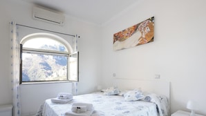 A bedroom with a bed, a window with a view of mountains, a painting on the wall, and a bedside table.
