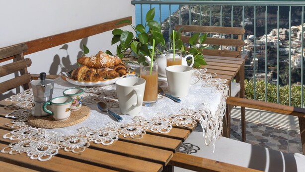A balcony with a wooden table set for breakfast, overlooking a coastal view.
