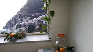 A window view of a coastal town with buildings, a mountain, and a table with oranges, a pitcher, and a bottle.