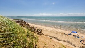 A beach with a wooden walkway leading to the water, a sandy beach area with a blue canopy, and a clear blue sky.