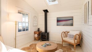 A cozy room with a round wooden table, a sofa, a wall-mounted painting, a window with blinds, and a wood-burning stove.