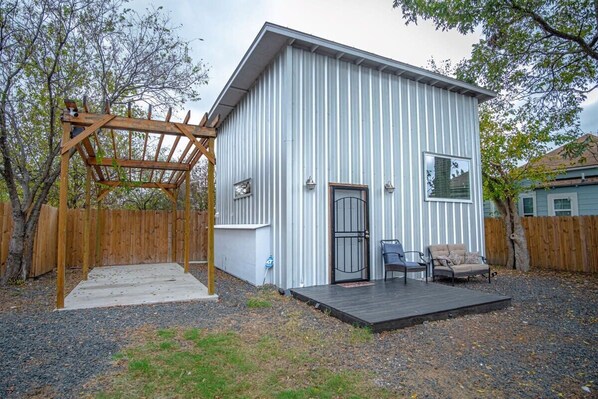 A small, single-story building with a metal exterior, a wooden pergola, and a small deck with two chairs. - Guest House with Loft Near Downtown (San Antonio)