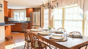 A dining area with a wooden table set for four, surrounded by wooden chairs. A chandelier hangs above the table, and there is a window with curtains on the right. - Walk to Water Hot Tub & Game Room! (Eastham)