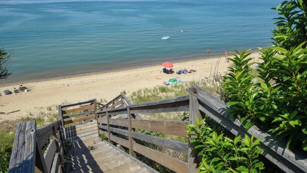 A wooden staircase leading to a beach with clear blue water, a sandy shore, and some greenery. - Walk to Water Hot Tub & Game Room! (Eastham)