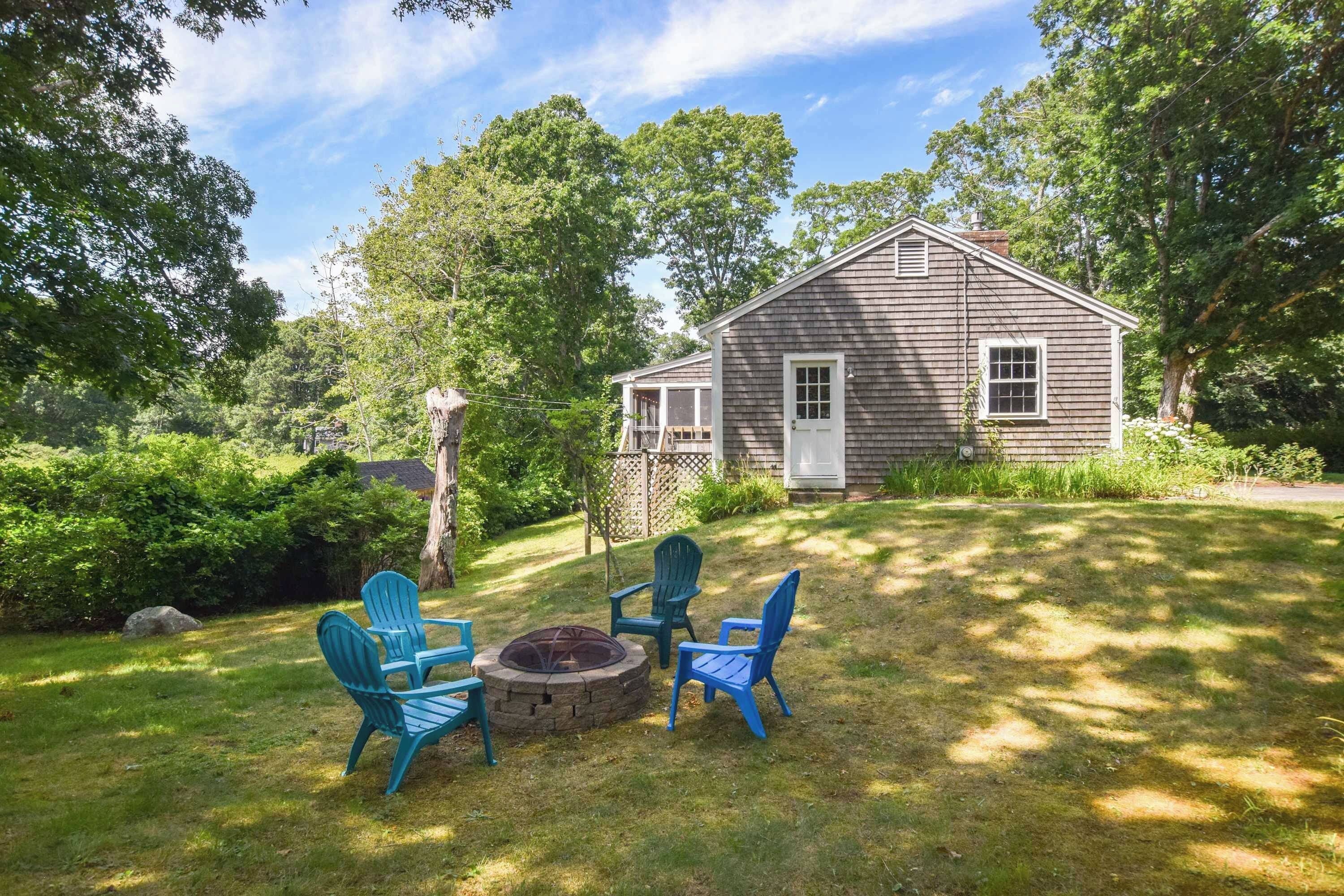 A wooden cabin with a fire pit and three Adirondack chairs in a grassy yard.
