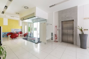 A modern lobby with a glass door entrance, a reception area with red chairs, a large potted plant, and a wall-mounted elevator. - Silver Hotel -  Matrimoniale (Matino)