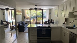 A modern kitchen with a central island, a dining area with a sofa, and a view of a garden through large windows. - Relax and Rejuvenate in the Heart of Kona. Minutes walk to town! (Kailua-Kona)