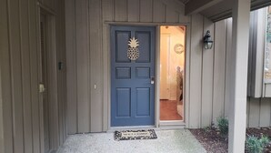 Une maison en bois avec une porte bleue ornée d’un motif représentant un ananas, un paillasson et un petit jardin avec des plantes.