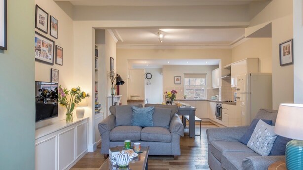 A living room with two sofas, a coffee table, and a TV. There is a kitchen in the background with a fridge and a microwave.