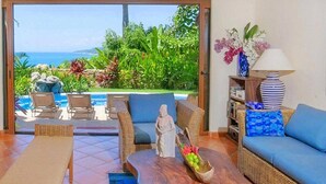 A room with a view of the ocean, wicker furniture, a statue, a wooden coffee table with fruit, and a lamp.
