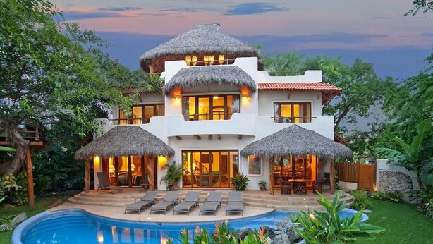 A two-story house with a swimming pool, thatched-roof structures, and lush greenery.