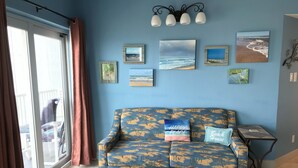 A room with a blue wall, a sofa with tropical prints, and several framed pictures depicting beach scenes. -  Cozy Beach Condo Awaits on Padre Island! (South Padre Island)