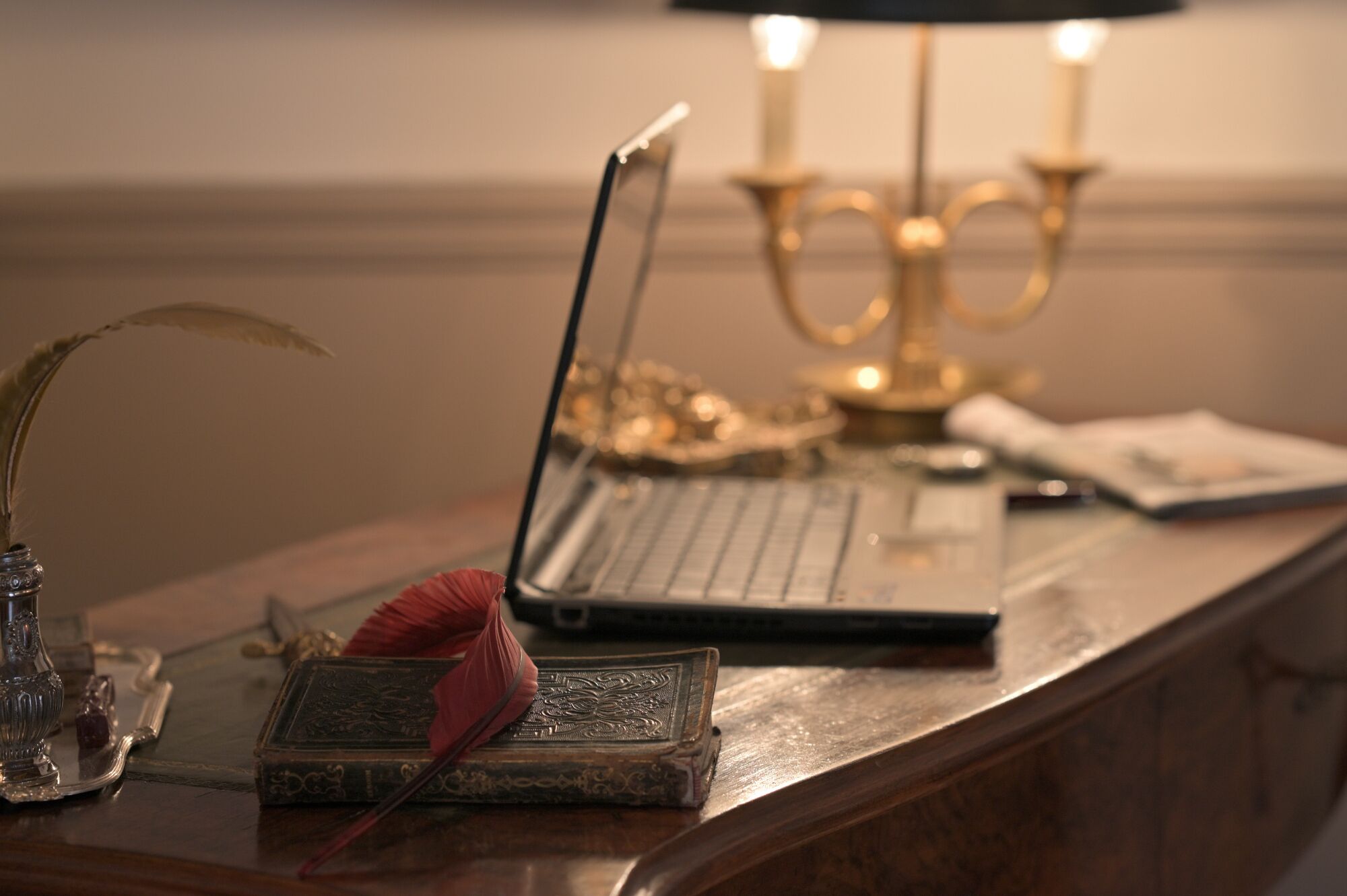 Un ordinateur portable posé sur un bureau en bois, avec une lampe décorative, une plume d’oie et un livre à la couverture ornée.