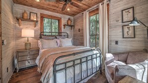 A rustic bedroom with a metal bed frame, wooden walls, and a large window offering a view of the forest.