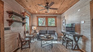 A cozy cabin interior with a wooden ceiling, a ceiling fan, a sofa, a coffee table, a TV, and a fireplace.