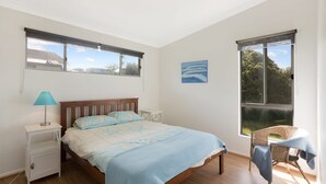 Quarto com cama, mesa de cabeceira, luminária, cadeira e duas janelas.