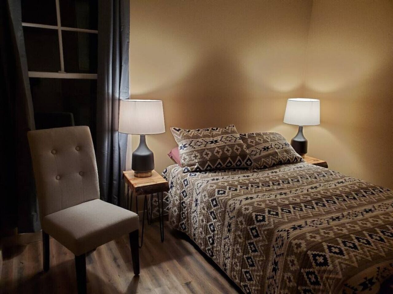 A hotel room with a bed, two bedside tables with lamps, a chair, and a window with curtains.